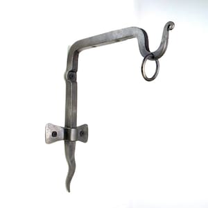 May include: A silver metal wall-mounted bracket with a decorative hook and a ring. The bracket is designed to hold a plant or other object.