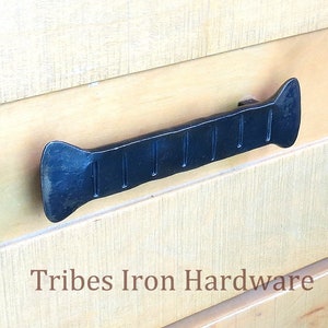 Könnte beinhalten: Schwarzer Metall-Schubladengriff mit einer strukturierten, geriffelten Oberfläche. Der Griff ist auf einer hellholzfarbenen Oberfläche montiert. Tribes Iron Hardware.