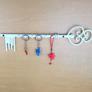 May include: White key-shaped wall mounted key holder with five hooks. The key holder has a decorative design with a heart shape and swirls. There are three keychains hanging from the hooks, one is red, one is blue, and one is red and white.