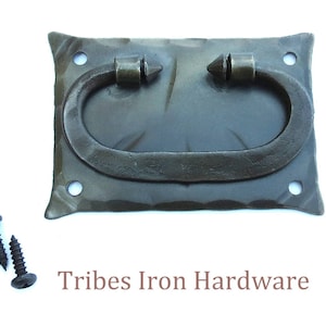 May include: A dark gray, hand-forged iron drawer pull with a rectangular backplate and a curved handle. The handle is attached with two conical rivets. Includes black screws. The text "Tribes Iron Hardware" is below the pull.