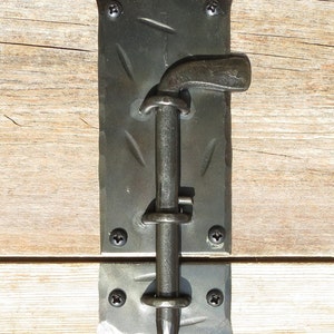 Puede incluir: Pestillo de puerta de metal negro con una superficie texturizada y un mango curvo. El pestillo est&aacute; fijado a una superficie de madera con tornillos.