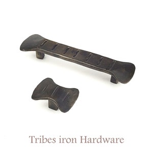 May include: Two rustic iron cabinet pulls with a hammered finish. The larger pull is rectangular with a slightly curved shape and the smaller pull is a rounded, symmetrical shape. Both pulls have a series of grooves running along the length of the pull. Tribes iron Hardware.