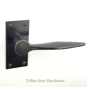 Könnte beinhalten: Schwarzer Eisentürgriff mit quadratischer Rückplatte und einem flachen, leicht gebogenen Hebel. Der Griff wird von Tribes Iron Hardware hergestellt.