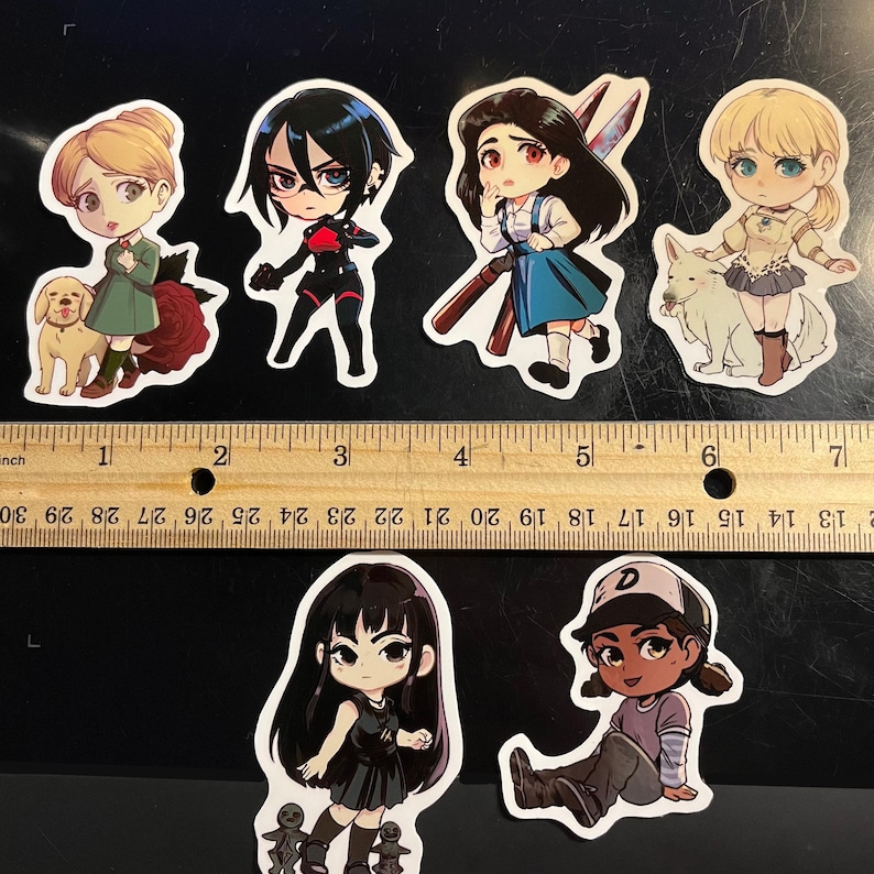 May include: Six die-cut vinyl stickers featuring chibi-style characters, including a blonde with a dog, a dark-haired figure in black, a character with scissors, a blonde with a white dog, a girl in a black dress, and a character in a cap, shown with a ruler.