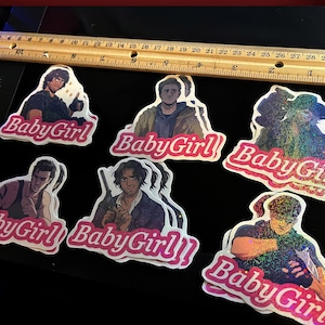 May include: A selection of stickers with character illustrations and the text "BabyGirl" in pink cursive. The stickers are displayed on a black surface, with a wooden ruler in the background. The ruler shows measurements in inches.