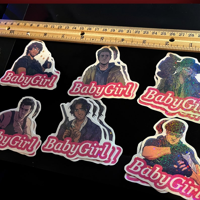 May include: A selection of stickers with character illustrations and the text "BabyGirl" in pink cursive. The stickers are displayed on a black surface, with a wooden ruler in the background. The ruler shows measurements in inches.