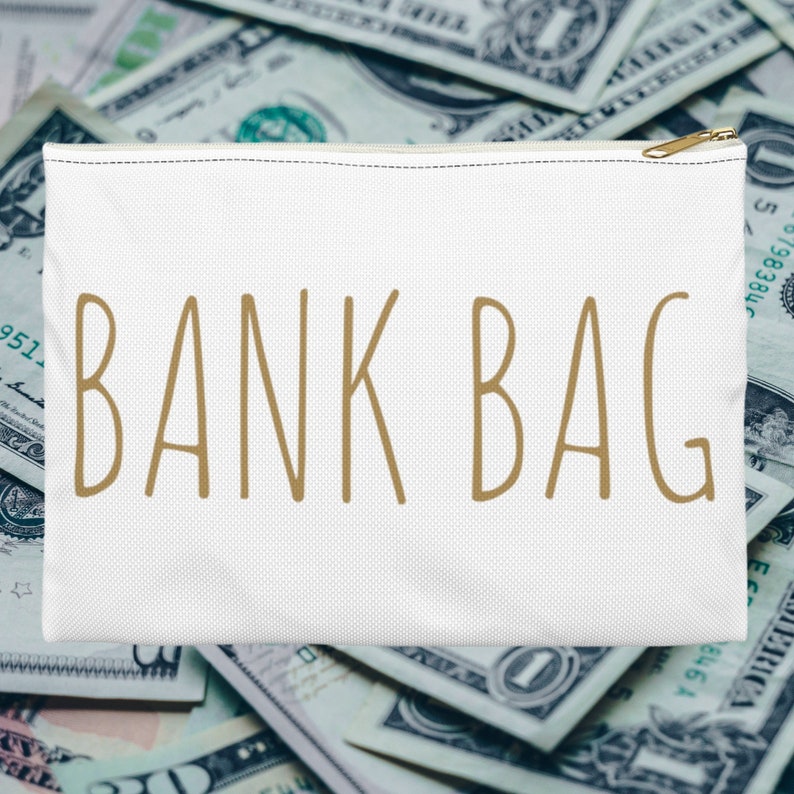 May include: White canvas zippered pouch with the text "BANK BAG" in gold lettering, sitting on a pile of US dollar bills.