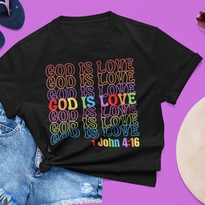 Könnte beinhalten: Schwarzes T-Shirt mit dem Spruch "GOD IS LOVE", der in Regenbogenfarben wiederholt wird. Der Text "1 Johannes 4:16" ist unter dem Regenbogentext gedruckt.