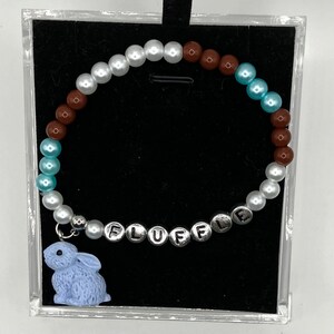 May include: A white, blue, and brown beaded bracelet with a silver charm that says "FLUFFLE" and a light blue bunny charm.