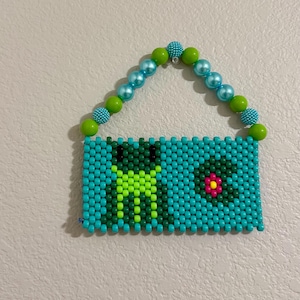 May include: A beaded frog-themed wall hanging. The rectangular body is turquoise, with a green frog and a pink flower. The handle is made of blue and green beads. A unique, handmade piece.
