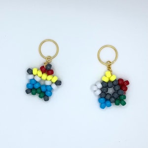 May include: Two star-shaped beaded keychains. Each keychain features a gold-colored ring and a star made of colorful beads. The beads are in various colors, including red, yellow, blue, gray, and green. The keychains are on a white background.
