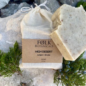 May include: A bar of soap and a muslin bag with a brown label. The label reads "FOLK BOTANICALS HIGH DESERT Juniper + Smoke SOAP 4.5 oz." The soap is light beige with dark specks. The bag is tied with a white string.