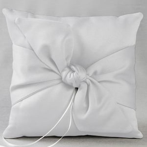May include: A square, white satin ring pillow. The pillow features a large, knotted bow made from the same satin material. Two long, thin satin ribbons extend from the knot. The pillow is designed for a wedding ceremony.