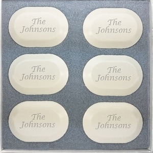 May include: A set of six ivory-colored, oval-shaped soaps, each inscribed with "The Johnsons" in a delicate font. The soaps are neatly arranged in a gray box, creating a personalized gift.