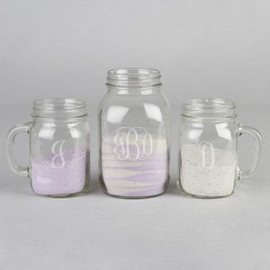 May include: Three glass mason jars with handles. The jars are filled with sand and have a monogram etched on the front. The jars are arranged in a row from left to right, with the largest jar in the middle.