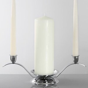 May include: A silver candle holder with three white candles. The center candle is thick and cylindrical, flanked by two thinner, tapered candles. The holder has a reflective surface.