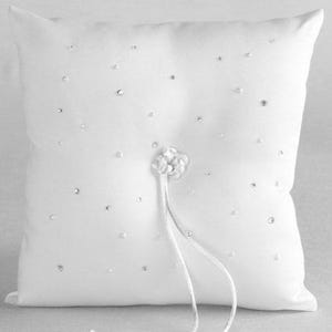 May include: A square, white ring bearer pillow. The pillow is decorated with scattered rhinestones and a central floral embellishment. White satin ribbons extend from the floral detail. A classic accessory for a wedding ceremony.