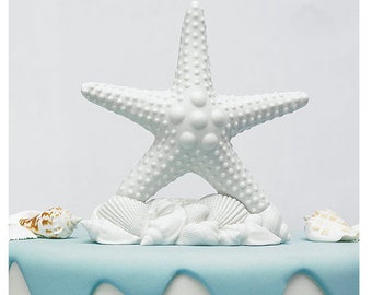 May include: A white starfish cake topper with a textured surface, resting on a bed of white seashells. The topper is placed on a cake with a light blue frosting design, evoking a beach theme.