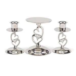 May include: A set of three silver candle holders with a heart design. The center holder is taller and has a round top for a cake or other centerpiece. Each holder has a small crystal in the center of the heart.