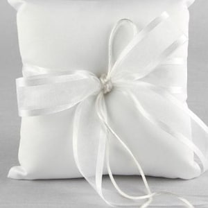 May include: A square, white satin ring pillow with a sheer white ribbon tied in a bow. The pillow is designed to hold wedding rings. The bow is centered on the pillow and has long, flowing ribbon ends.