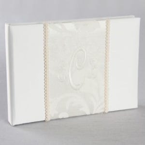 May include: A white photo album with a decorative ivory fabric panel featuring an embroidered letter "C". The panel is bordered with delicate lace trim. The album is rectangular and appears to be new.
