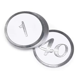 May include: A stack of circular white tokens with a metallic silver border and the number 1 embossed in silver on the top piece, accompanied by a single separate circular white token featuring the number 40 in a metallic silver finish.