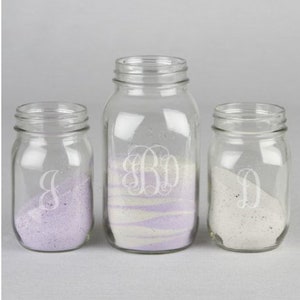 May include: Three clear glass jars with lids, filled with different colored sand. The jars are decorated with white, engraved monograms: J, BD, and D.
