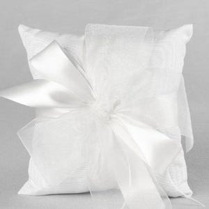 May include: A square, white ring bearer pillow with a textured surface. A large, white satin bow and sheer ribbon overlay the pillow, creating a layered effect. The pillow is designed for a wedding ceremony.
