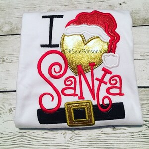 May include: White t-shirt with the embroidered phrase "I Santa". The design includes a red and gold Santa hat, a gold heart, and a black belt with a gold buckle. The text "OhSewPersonalized" is also visible.