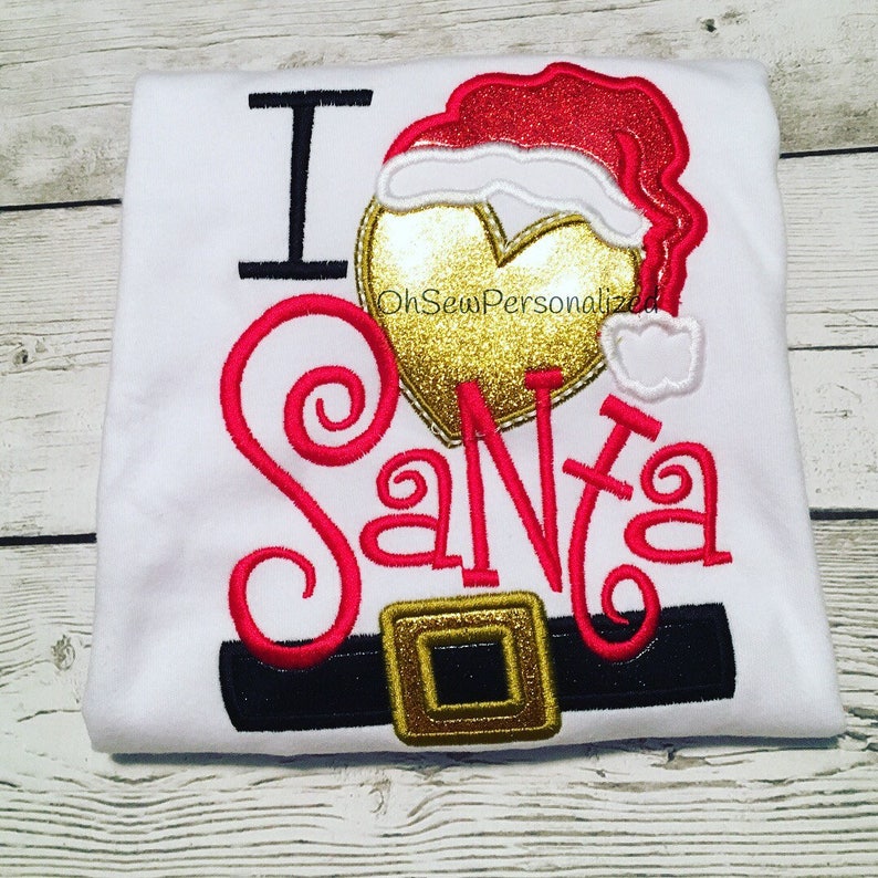 May include: White t-shirt with the embroidered phrase "I Santa". The design includes a red and gold Santa hat, a gold heart, and a black belt with a gold buckle. The text "OhSewPersonalized" is also visible.