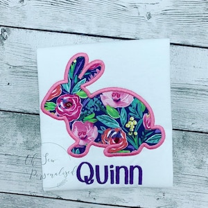 May include: White t-shirt featuring a floral bunny design with a pink outline. The bunny is filled with pink roses, green leaves, and other flowers on a dark blue background. The name "Quinn" is embroidered in purple.