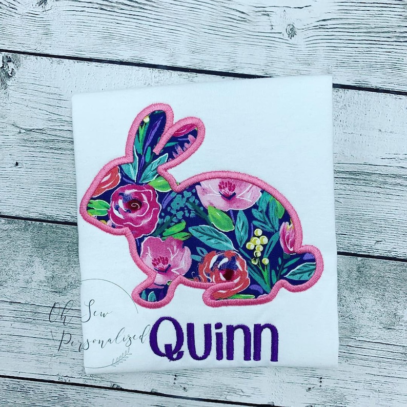 May include: White t-shirt featuring a floral bunny design with a pink outline. The bunny is filled with pink roses, green leaves, and other flowers on a dark blue background. The name "Quinn" is embroidered in purple.