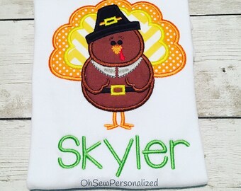 May include: White t-shirt featuring an embroidered turkey wearing a pilgrim hat, with the name "Skyler" in green script. The turkey has a brown body, orange and yellow accents, and a red wattle. The text "OhSewPersonalized" is at the bottom.