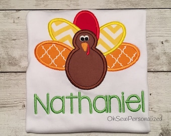 May include: White t-shirt with a colorful turkey applique. The turkey has a brown body, red wattle, and patterned feathers in orange, yellow, and red. The name "Nathaniel" is embroidered in green script. The text "OhSewPersonalized" is also visible.