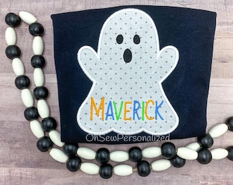 May include: A navy blue shirt with a white ghost applique featuring a cross pattern. The name "MAVERICK" is embroidered in colorful thread. A black and white beaded garland frames the shirt. The text "OhSewPersonalized" is at the bottom.