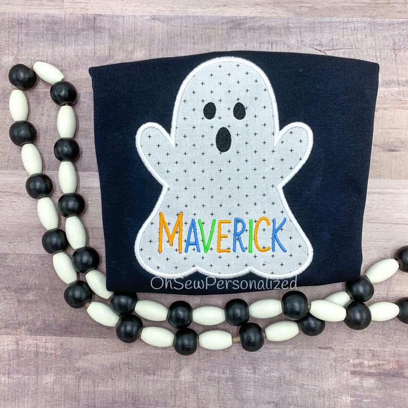 Op de afbeelding: Een marineblauw shirt met een witte spookapplicatie met een kruispatroon. De naam "MAVERICK" is geborduurd met gekleurd garen. Een zwart-witte kralenkrans omlijst het shirt. De tekst "OhSewPersonalized" staat onderaan.