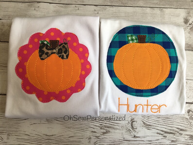 May include: Two white t-shirts with pumpkin appliqu&eacute;s. One has an orange pumpkin with a leopard print bow on a pink polka dot scalloped border. The other has an orange pumpkin on a blue and green plaid circle with the name "Hunter" stitched below.