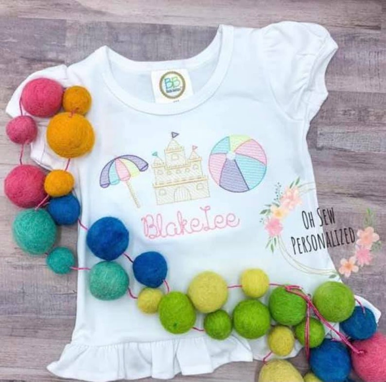 Puede incluir: Camiseta blanca de manga corta con un dise&ntilde;o de tem&aacute;tica playera, que incluye una sombrilla, un castillo de arena y un bal&oacute;n de playa. El nombre "Blake Lee" est&aacute; bordado en rosa. La camiseta est&aacute; decorada con una guirnalda de bolas de fieltro de colores. Tambi&eacute;n se ve el texto "Oh Sew Personalized".