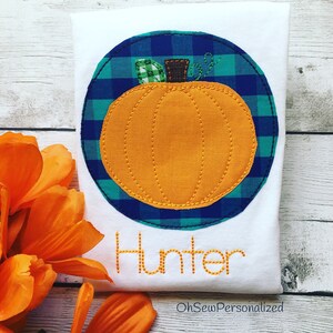 May include: White t-shirt with an orange pumpkin applique inside a blue and green plaid circle. The name "Hunter" is stitched in orange thread below the pumpkin. The text "OhSewPersonalized" is at the bottom.