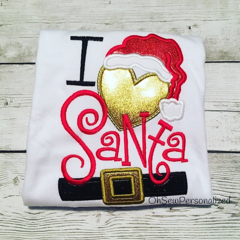 May include: White t-shirt with the text "I Santa" in red script. The design includes a gold heart, a Santa hat, and a black belt with a gold buckle. The shirt is folded on a wooden surface.