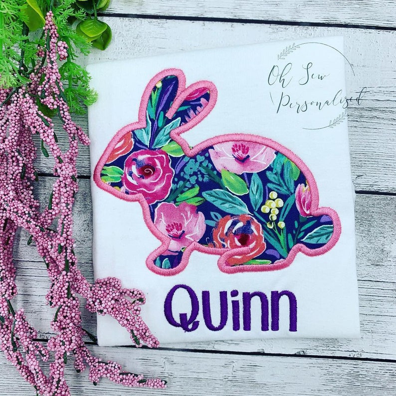 May include: White t-shirt featuring a floral bunny design with pink trim. The bunny is filled with pink and purple flowers on a navy blue background. The name "Quinn" is embroidered in purple below the bunny. The text "Oh Sew Personalized" is in the upper right corner.