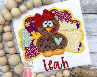 May include: White t-shirt with a turkey applique. The turkey has a brown body, a red bow, and a green heart. The name "Leah" is embroidered in red. The design is surrounded by a floral pattern in yellow, purple, and burgundy.