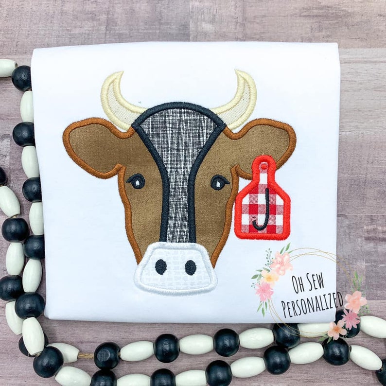May include: White t-shirt featuring an embroidered cow face. The cow has brown fur, a black and white plaid patch, and a red and white plaid ear tag with the letter "J". The shirt has the text "Oh Sew Personalized".