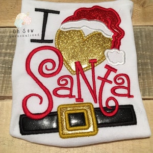 May include: White t-shirt with the embroidered phrase "I ❤️ Santa." The design includes a gold glitter heart, a red Santa hat, and a black belt with a gold buckle. The shirt is a festive holiday item.