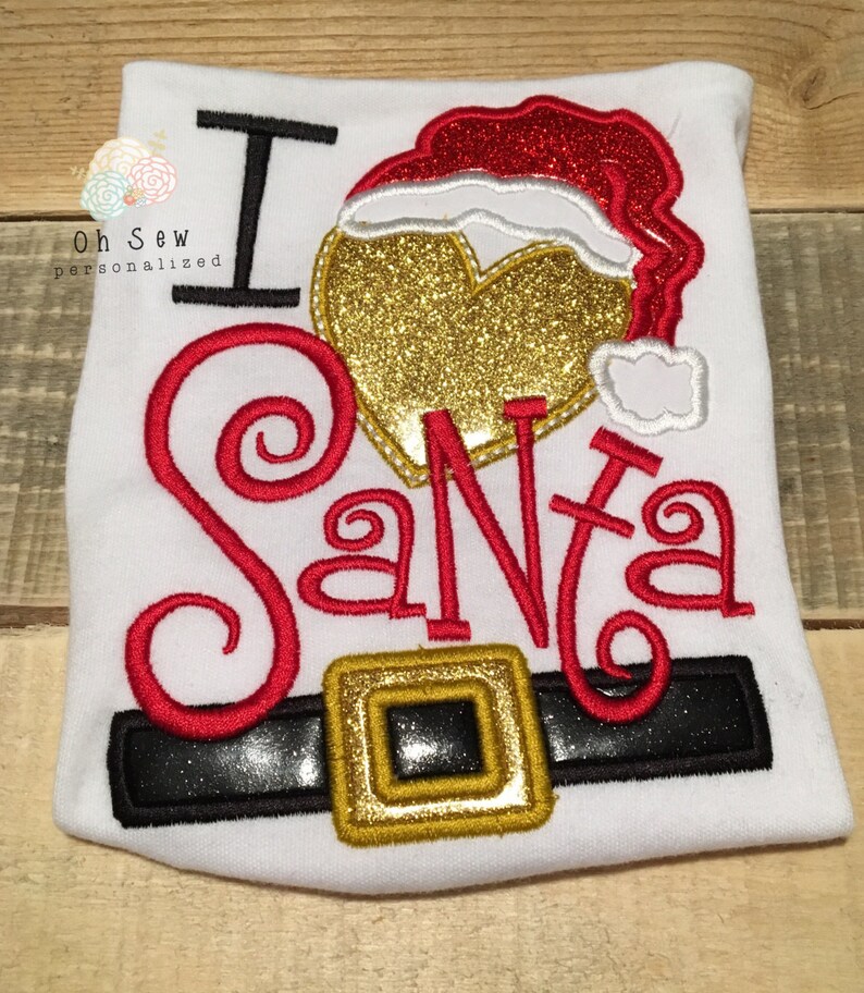 May include: White t-shirt with the embroidered phrase "I ❤️ Santa." The design includes a gold glitter heart, a red Santa hat, and a black belt with a gold buckle. The shirt is a festive holiday item.