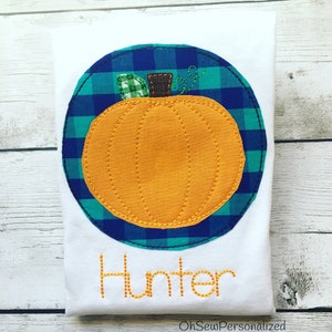 May include: White t-shirt featuring an orange pumpkin applique with a green and brown stem, set within a blue and green plaid circle. The name "Hunter" is embroidered in orange thread below the pumpkin. The design is perfect for fall.