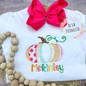 May include: White baby bodysuit with an embroidered pumpkin design in shades of orange, pink, and teal. The pumpkin has a brown stem and green vines. The name "McKinley" is embroidered in colorful letters. A large pink bow sits at the top.