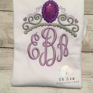 May include: White t-shirt with a purple embroidered tiara design and monogram. The tiara features a glittery purple center stone, gray accents, and small heart details. The monogram includes the letters EBA in a matching purple script. The text "Oh Sew personalized" is at the bottom.
