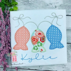 May include: White t-shirt with three fish-shaped appliqu&eacute;s in orange, floral, and blue patterns, each with a silver bow. The name "Kylie" is embroidered in blue script below the fish. The shirt is from Oh Sew Personalized Clothing.