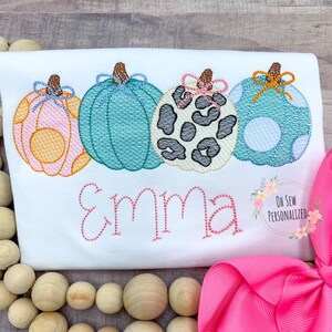 May include: White t-shirt featuring four embroidered pumpkins in pink, teal, and leopard print. The name "Emma" is stitched in pink below the pumpkins. A pink bow and wooden beads are in the background. The text "Oh Sew Personalized" is in the lower right corner.